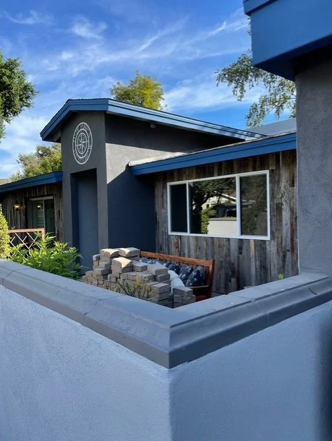 Modern home exterior with blue metal roof, gray stucco walls, and stone patio seating area