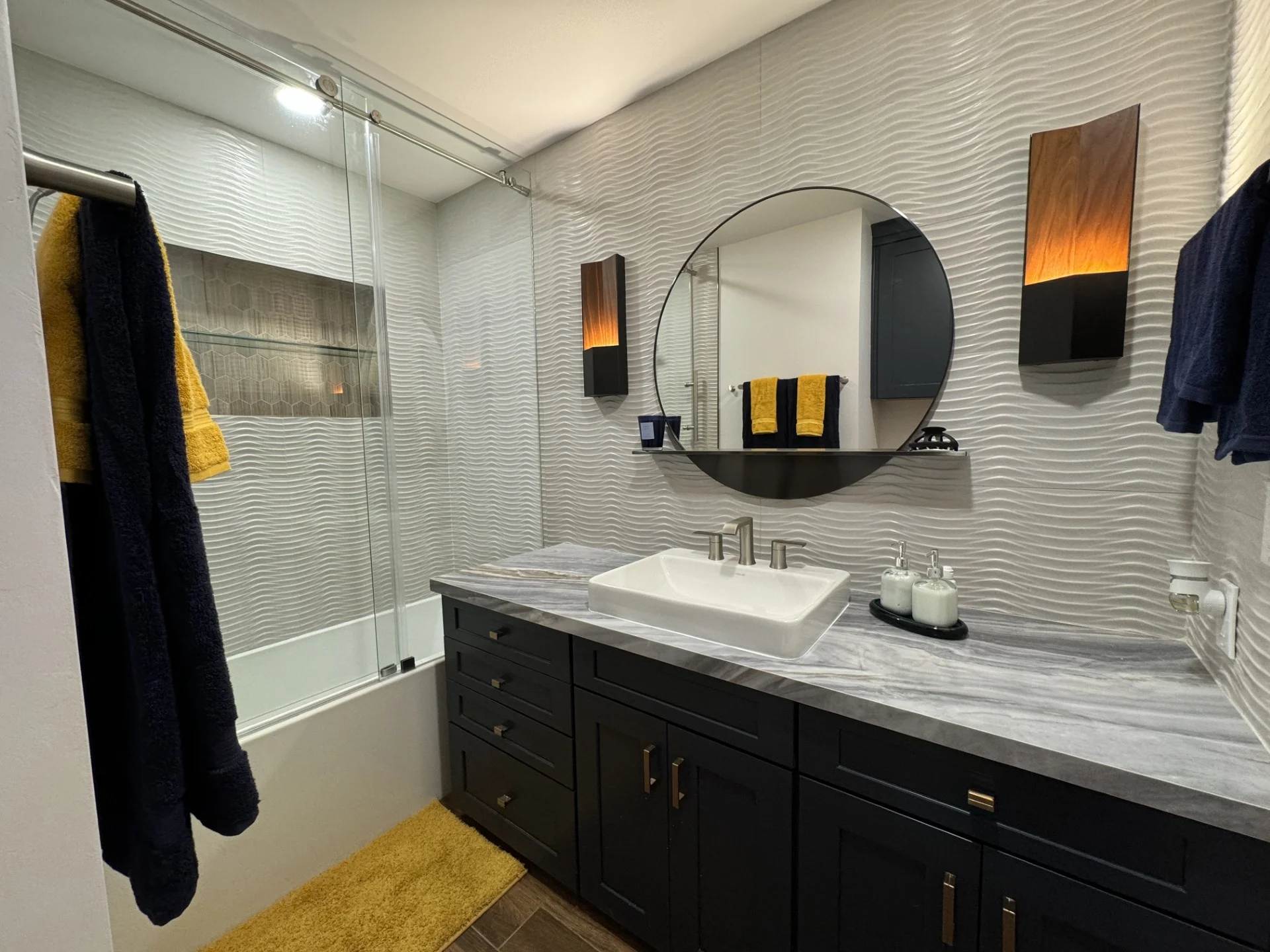 Modern bathroom with white textured tile walls, black vanity, marble countertop, and frameless glass shower
