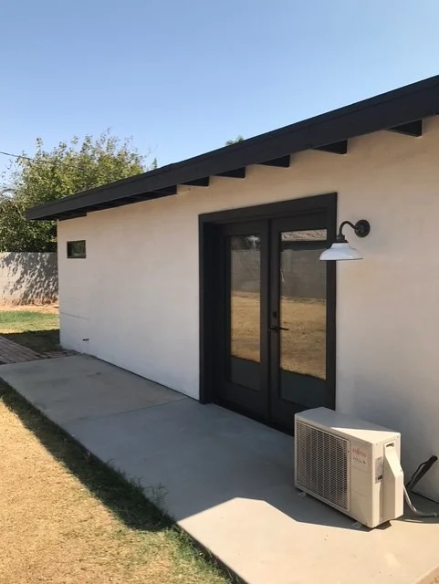 White stucco structure with black metal roof overhang, glass doors, and concrete patio
